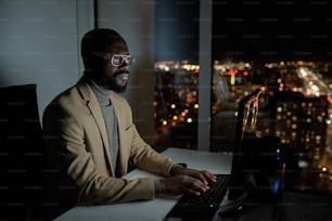 Oficinista africano barbudo en ropa formal escribiendo y mirando la pantalla de la computadora mientras trabaja horas extras por la noche