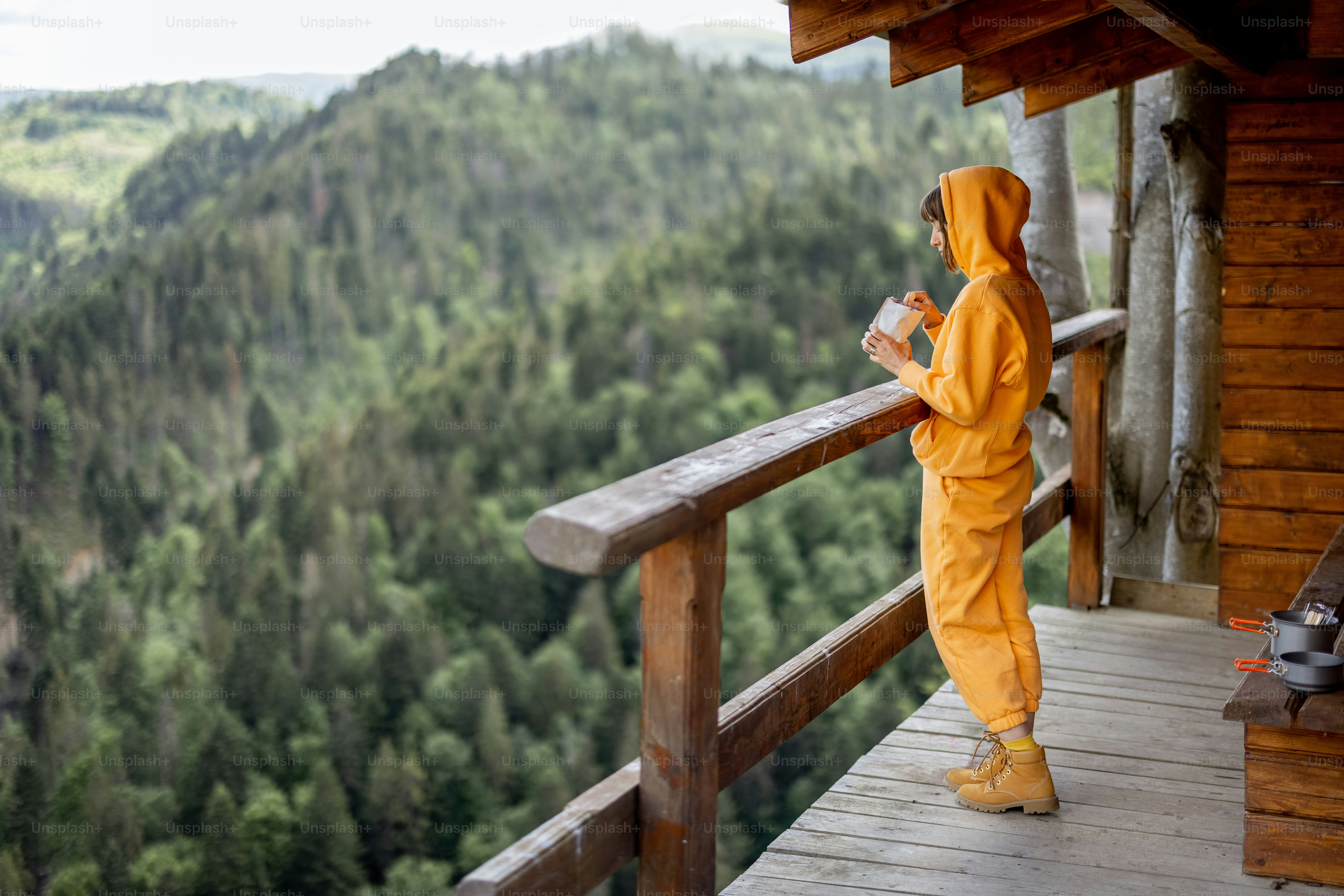 Young woman enjoys great mountain landscape and eats sublimated food for hiking, while standing on a wooden terrace of tiny house. Concept of solitude in nature and food for hiking