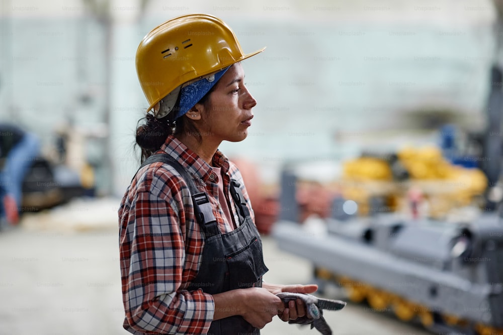 Side view of young tired female worker of factory in hardhat and coveralls standing in front of camera in large warehouse