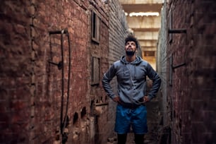 Portrait of focused motivated afro-american young handsome sportive man with earphones standing inside of the abandoned place in the middle of two walls and thinking while looking up.