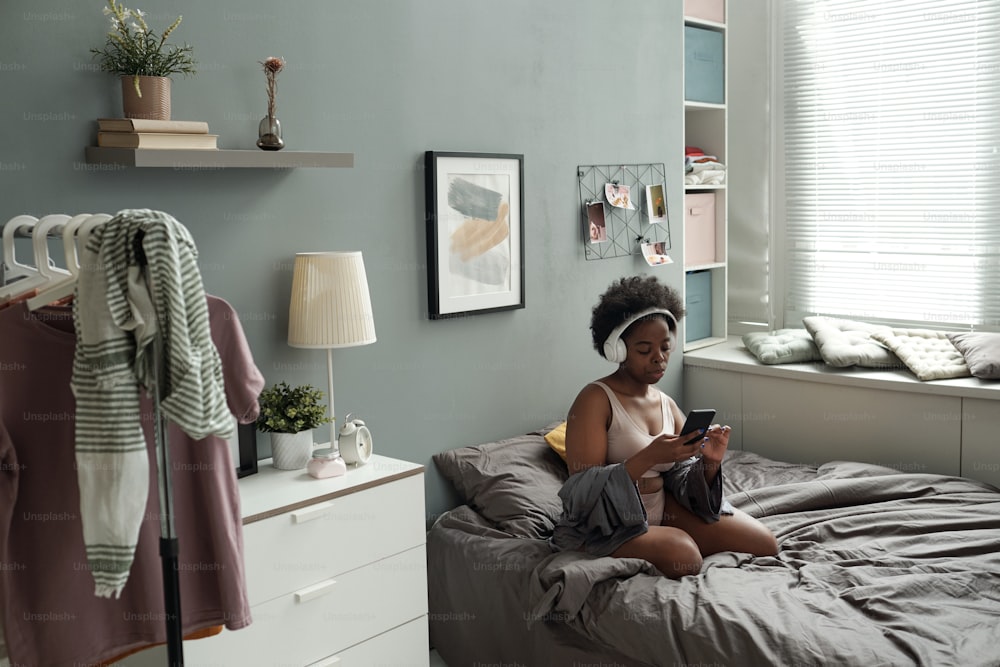 Young woman in headphones scrolling through playlist in smartphone while relaxing in bed