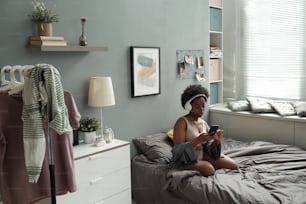 Giovane donna in cuffie che scorre la playlist nello smartphone mentre si rilassa a letto