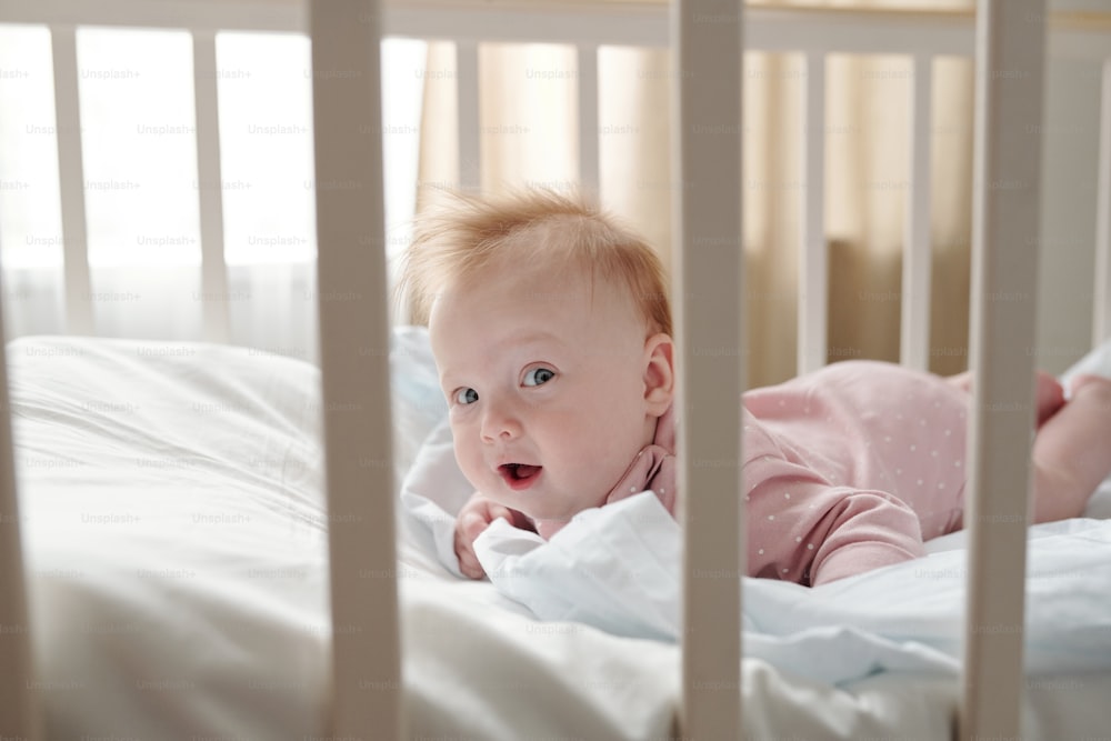 Happy baby girl looking at camera while creeping on white linen in her crib