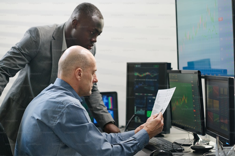Mature Caucasian and young Black men working together in stock trading company looking through document