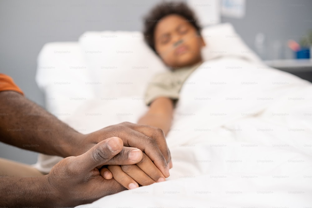 Close-up of father holding hand of his sick little son while he lying in bed at hospital