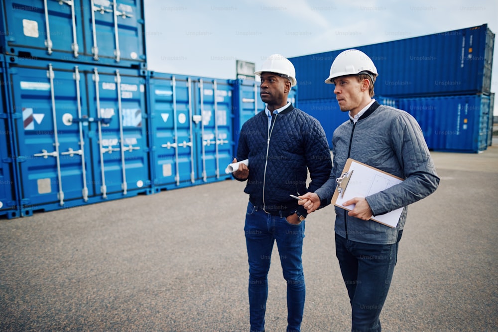 Two engineers wearing hardhats and talking together while standing in a freight yard full of shipping containers