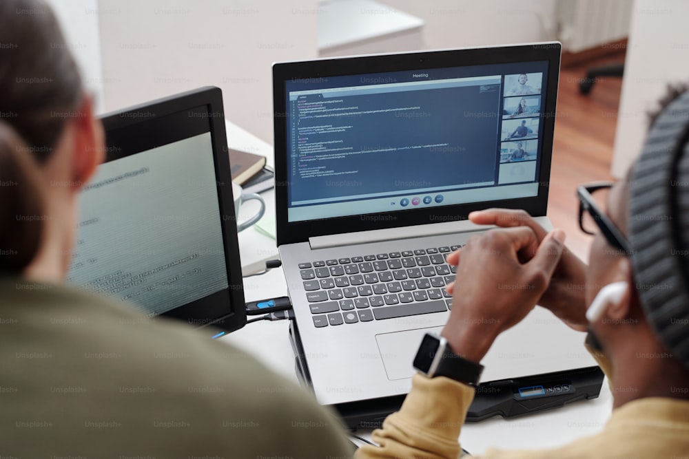 Mani di un giovane uomo nero sulla tastiera del laptop con video chat sullo schermo mentre è seduto sul posto di lavoro accanto alla collega femminile