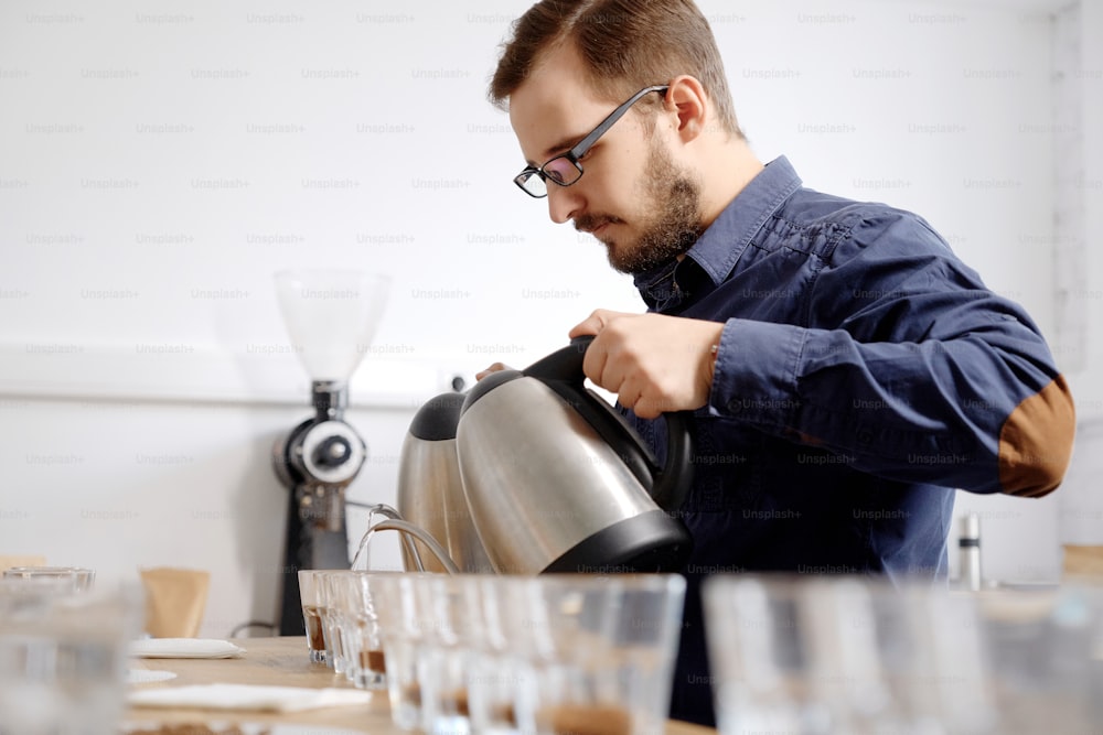 Barista masculino haciendo pucheros hirviendo agua en tazas de vidrio con café molido de dos hervidores, preparando café fresco para el examen de cata
