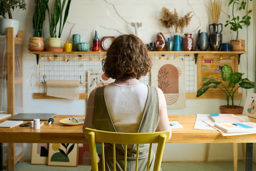 Rear view of young creative woman sitting by table in workshop with handmade souvenirs on shelf along wall and creating new paintings