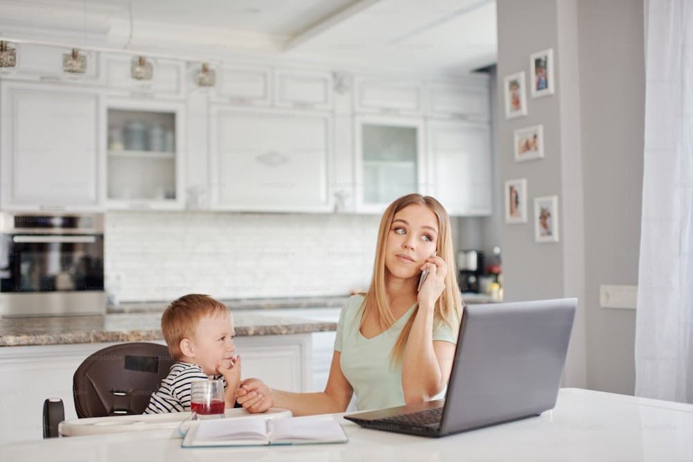 Portrait of mid age caucasian blond woman with child being busy on the phone and feeding the baby. They are at home, wearing pyjamas