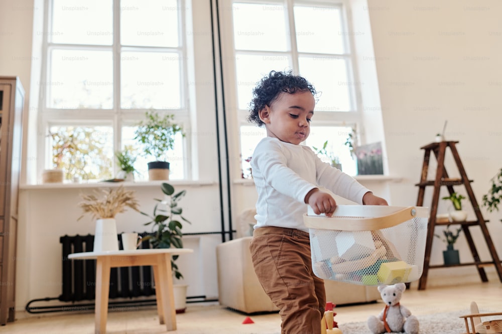 Serious independent little African-American boy with curly hair carrying box of toys across living room