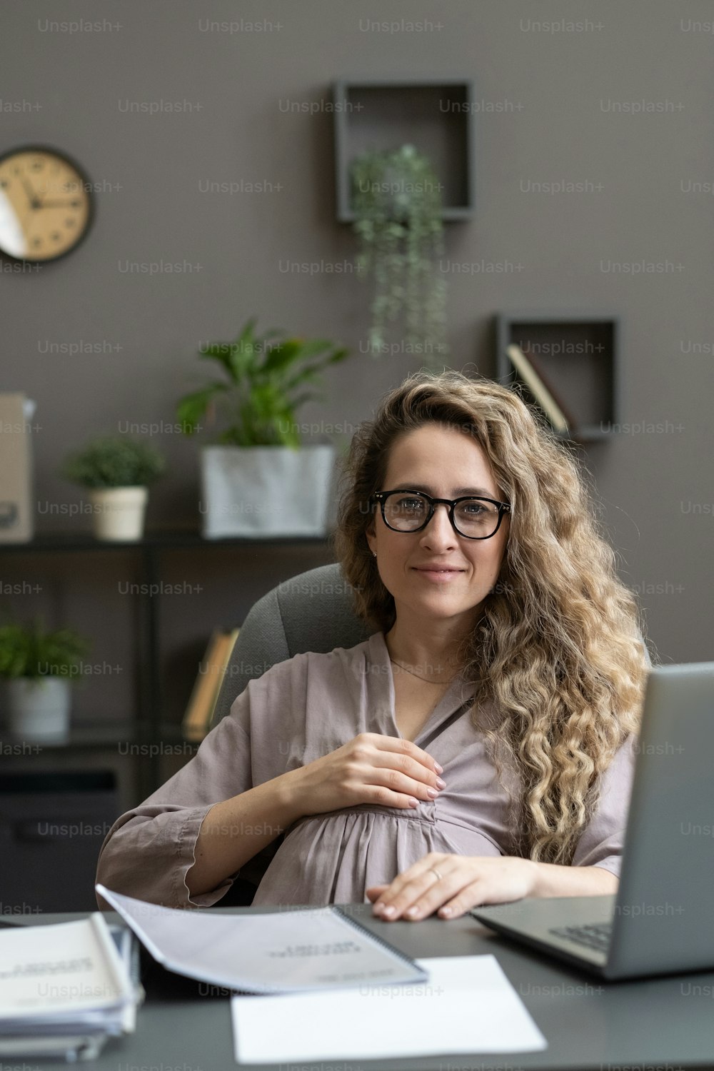 Portrait of young pregnant woman in eyeglasses looking at camera while sitting at her workplace with laptop at office