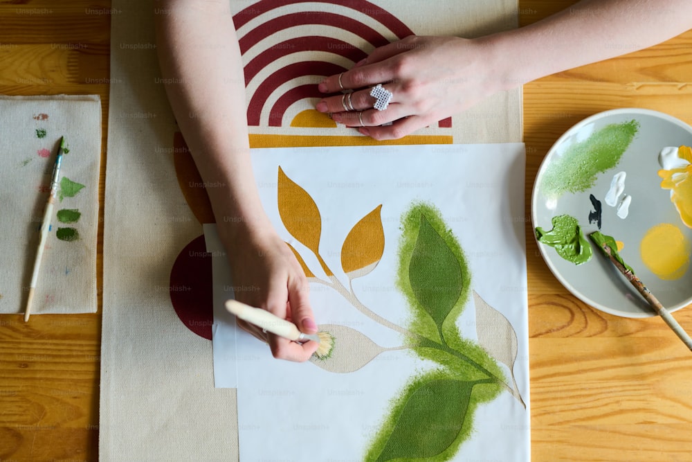 Above angle of hands of young female artist with paintbrush painting green leaves on paper while sitting by table in workshop