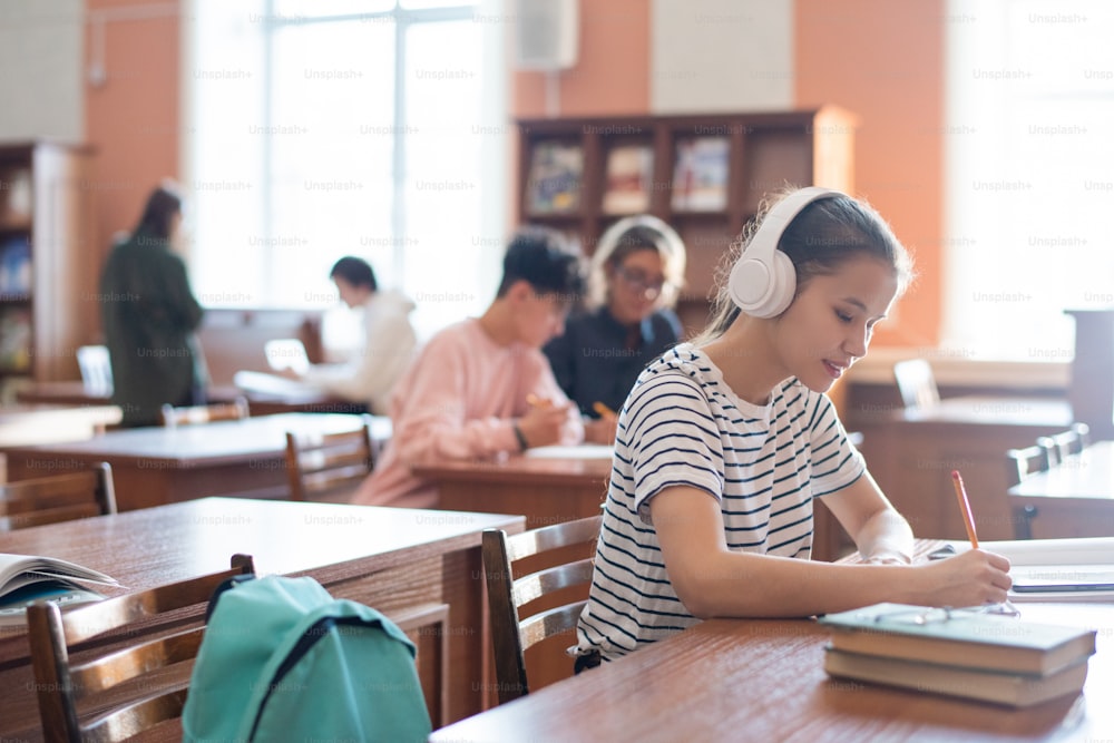 Estudiante universitario contemporáneo con auriculares que escribe el plan del seminario en el bloc de notas mientras está sentado en la biblioteca