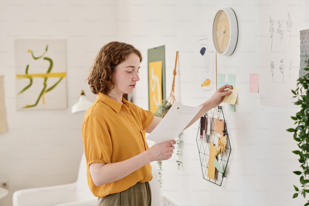 Young creative freelancer looking through information in paper document while standing in front of wall and sticking notepapers