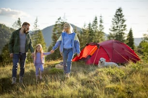 Young caucasian family with little girl walk together on green meadow while traveling with tent in the mountains. Happy family summer vacation at campsite