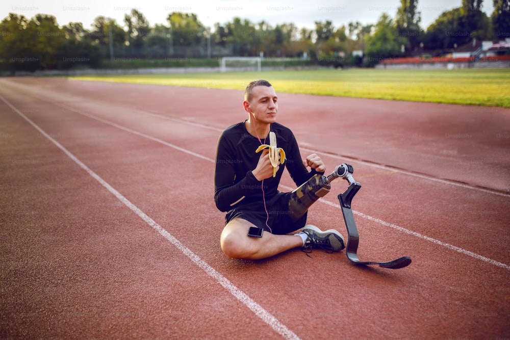 Guapo caucásico deportivo discapacitado joven en ropa deportiva y con pierna artificial sentado en la pista de carreras, escuchando música y comiendo plátano.