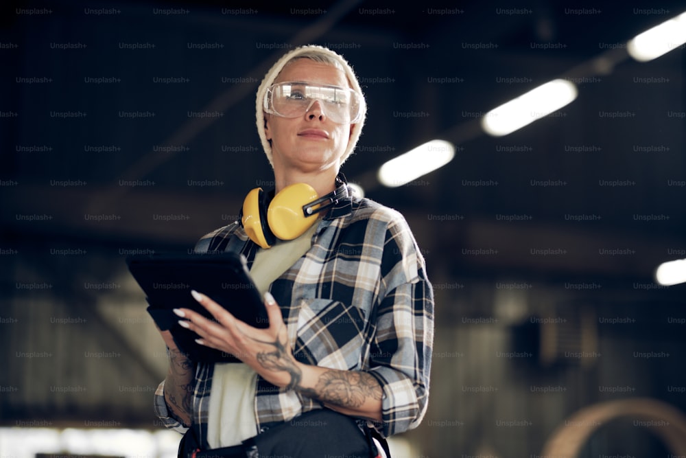 Portrait of blond woman in her 40s in protective eyewear, hat, and plaid shirt. She is holding digital tablet while working at factory
