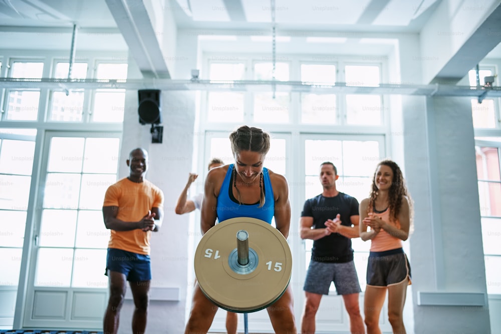 Group of young people cheering on their female friend exercising with weights while working out together in a gym
