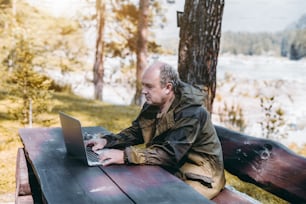 An adult partly bald game warden in the overalls of marsh color is sitting outdoor on the bench at the big wooden table and having a conversation with his family via video call from laptop