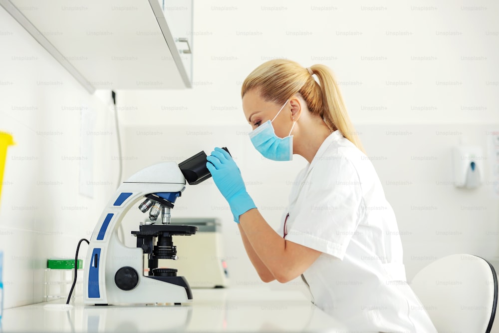 Beautiful lab assistant with rubber gloves and face mask on sitting in laboratory and looking trough microscope.