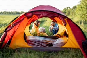 Giovane coppia prepara il cibo mentre viaggia con la tenda sulla natura. L'uomo e la donna trascorrono l'estate con il loro cane in campeggio. Vista attraverso la porta della tenda