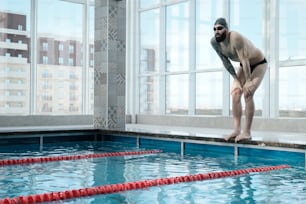 Fit bearded man in swimming goggles and cap standing on edge of poolside and preparing for swimming