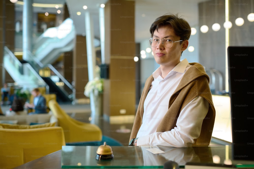 Young Asian man in casualwear and eyeglasses waiting for receptionist while standing by counter and looking at camera in hotel lounge