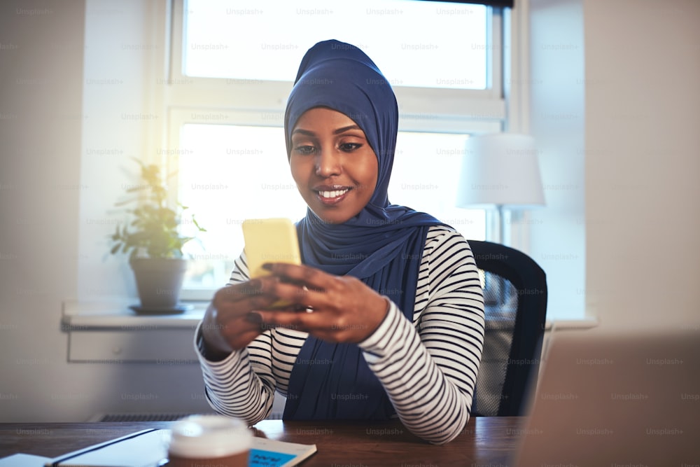 히잡을 쓴 젊은 무슬림 여성 기업가가 홈 오피스의 테이블에서 일하는 동안 문자 메시지를 보내고 있다