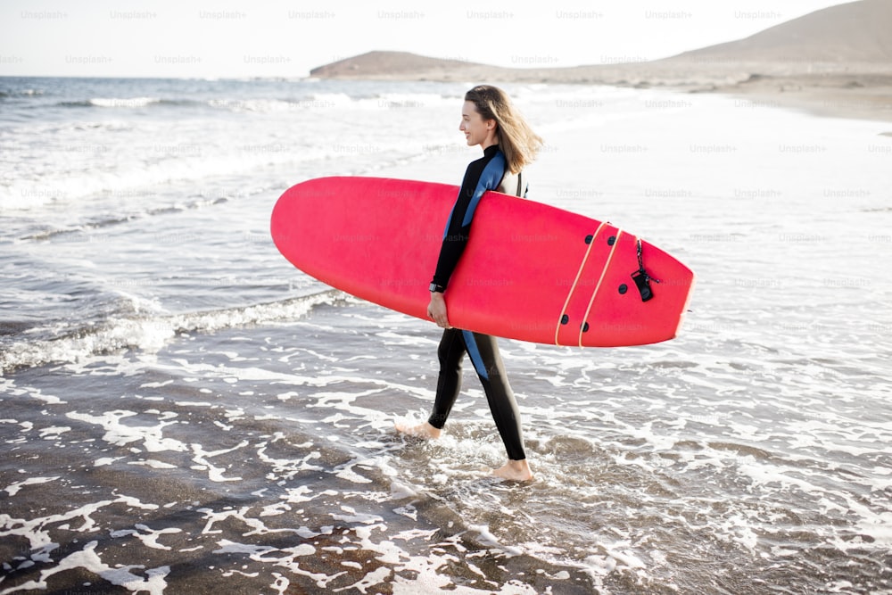 Mulher nova em roupa de mergulho caminhando com prancha de surf na bela praia selvagem em um pôr do sol. Conceito de desporto aquático e estilo de vida ativo