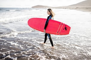 Young woman in wetsuit walking with surfboard on the beautiful wild beach on a sunset. Water sport and active lifestyle concept