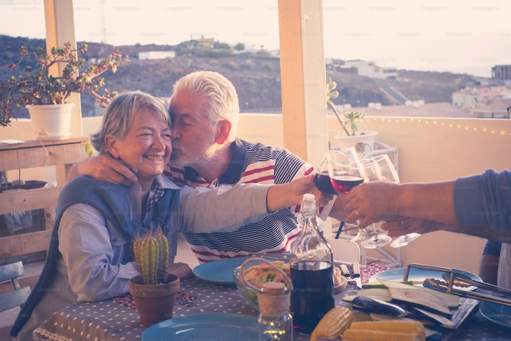 Gruppe von älteren älteren Freunden Erwachsene essen und feiern schöne Zeit auf der Dachterrasse im Freien mit Wein und Essen. Spaß haben und sich während des Sonnenuntergangs mit wunderschöner Sonnenbeleuchtung und herrlichem Blick auf das Meer und andere Dächer küssen.