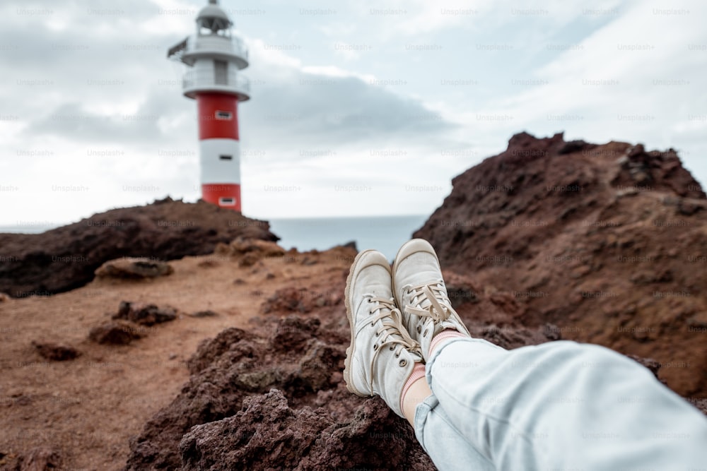 Young traveler resting on the rocky land, traveling volcanic landscapes on the ocean shore near the lighthouse. cropped view focused on the woman's shoes