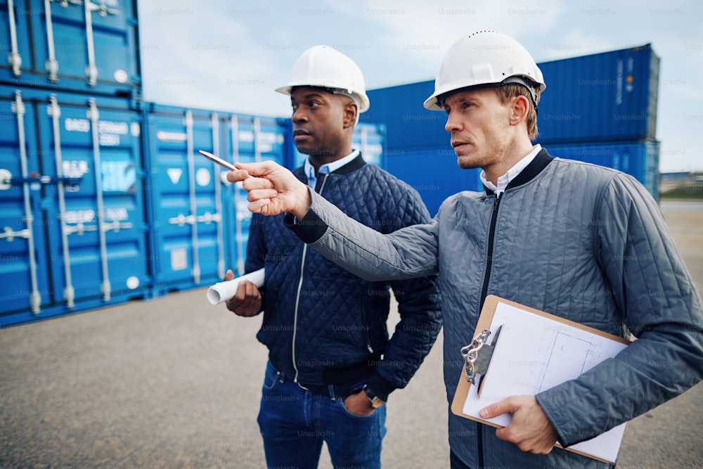 Dois engenheiros usando capacetes conversando juntos enquanto estão em pé em uma grande doca de transporte comercial rastreando o estoque