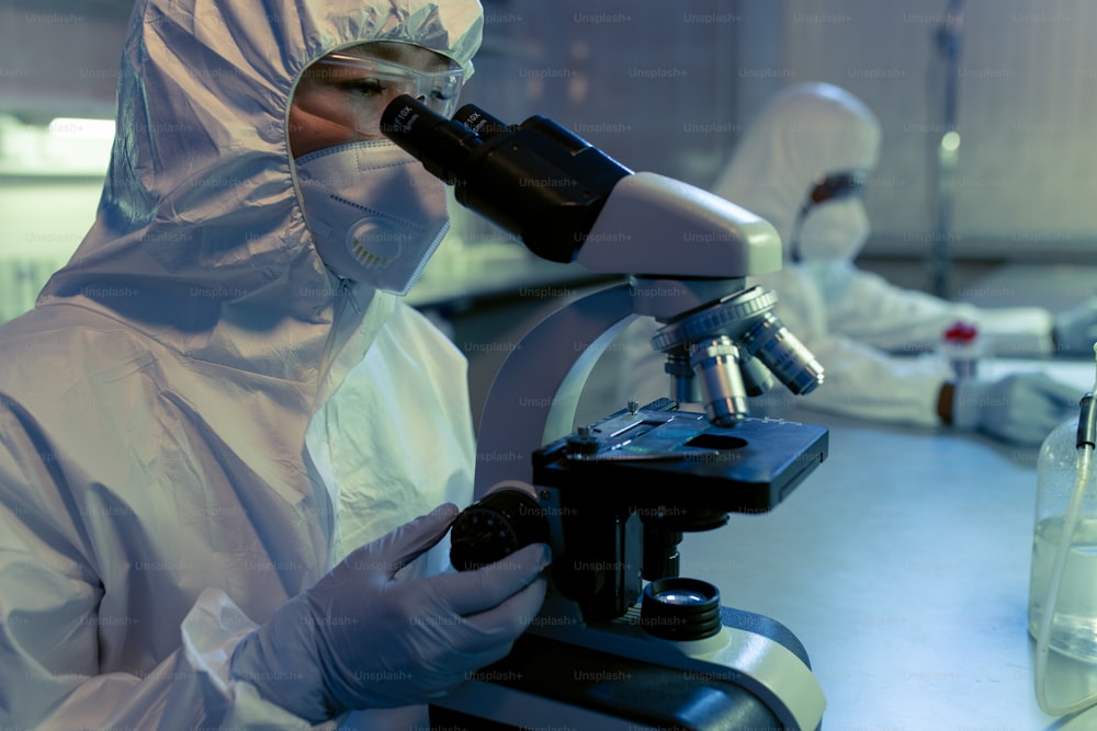 Química con ropa de trabajo protectora mirando a través del microscopio y examinando muestras durante un experimento científico en el laboratorio