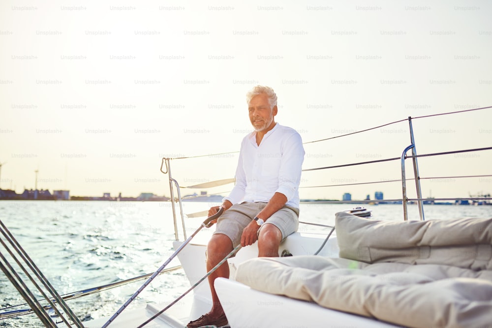 Homem maduro sentado no convés de um barco enquanto sai para uma vela ao longo da costa em um dia ensolarado