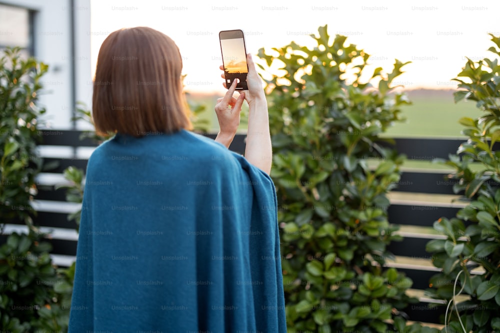 Frau in Karo macht Foto am Telefon und genießt den wunderschönen Sonnenuntergang im Hinterhof ihres Landhauses. Freizeit im Garten verbringen
