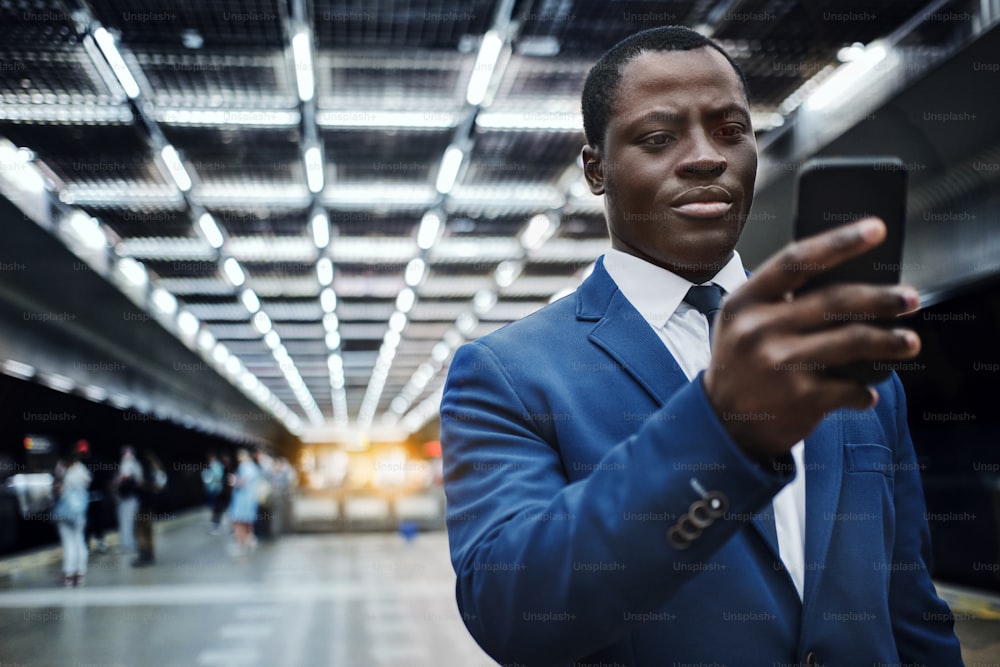 Retrato de un hombre negro confiado que viste traje azul y corbata, usando un teléfono inteligente en la estación de metro para navegar