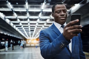 Portrait of confident black man wearing blue suit and tie, using smart phone at subway station to navigate
