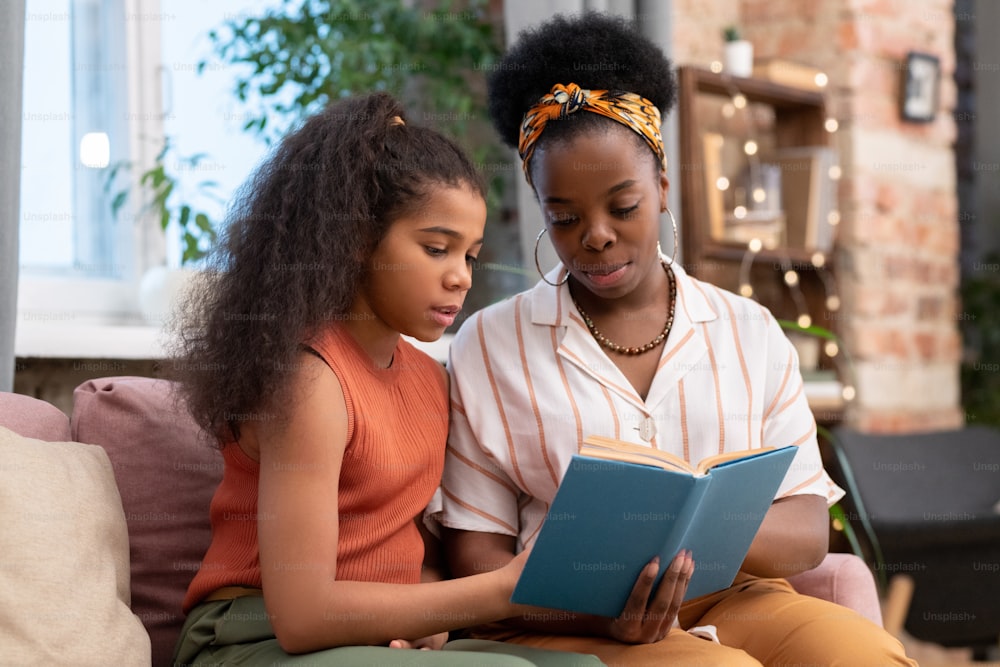 Mujer joven de etnia africana y su linda hija adolescente sentada en el sofá y leyendo un libro mientras se prepara para la lección de literatura