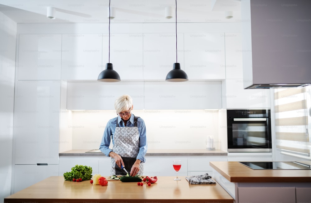 Gorgeous caucasian senior woman in apron shopping cucumber and making salad while standing in kitchen. On kitchen counter are vegetables and glass of wine.