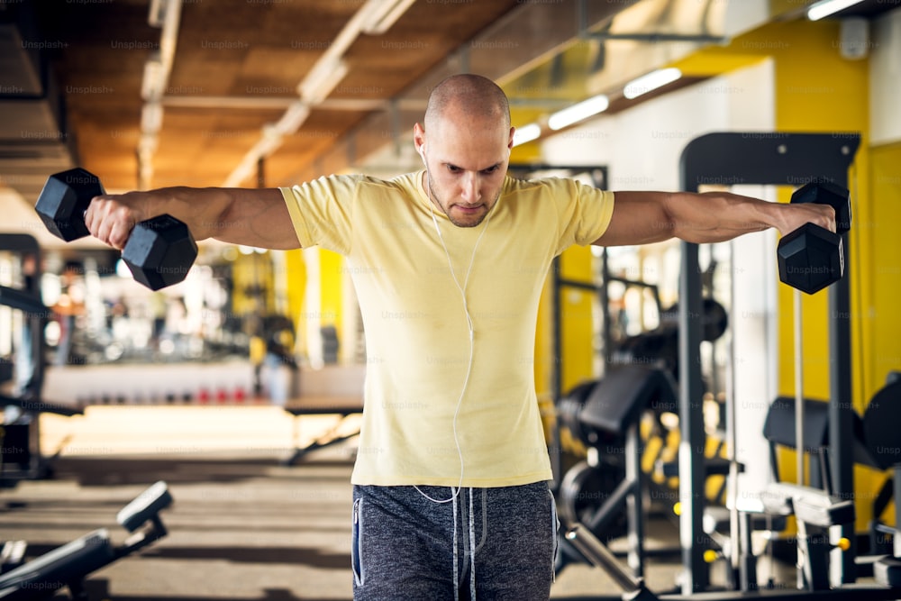 Portrait of strong muscular active healthy young man raising dumbbells with open arms and listening music in the gym.