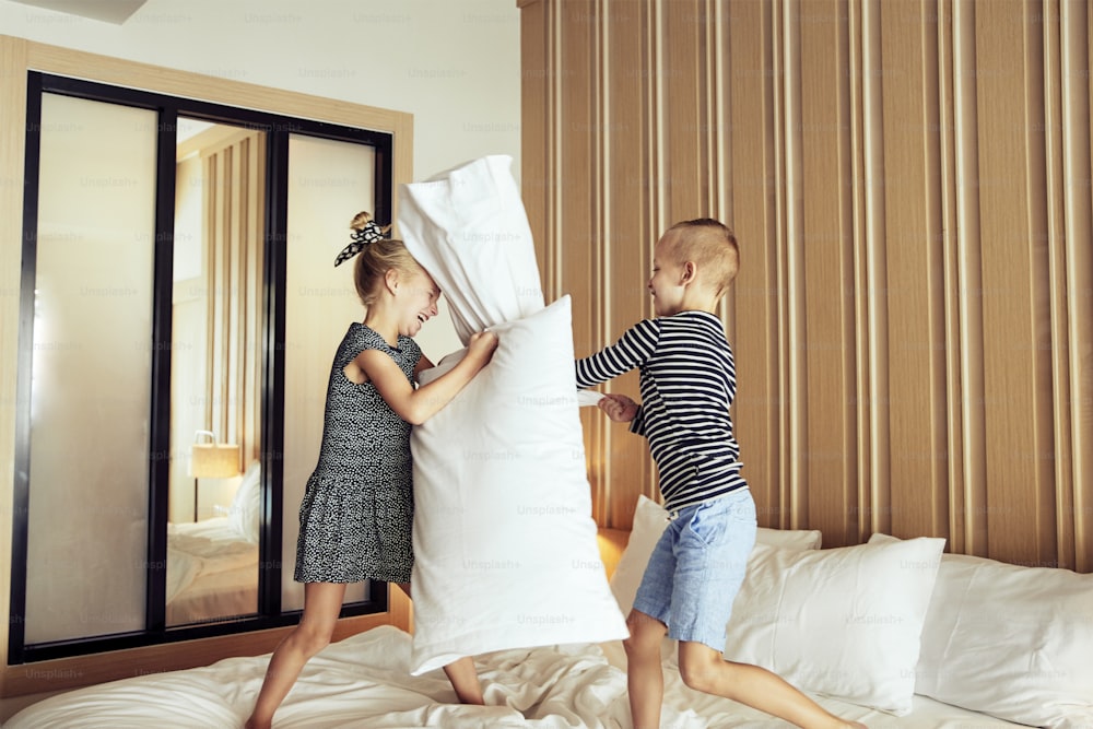 Cute little brother and sister laughing while having a pillow fight together on their bed at home
