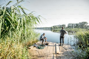 아침에 호수에 녹색 갈대가 있는 아름다운 나무 부두에서 피크닉을 즐기는 동안 휴식을 취하는 두 명의 어부