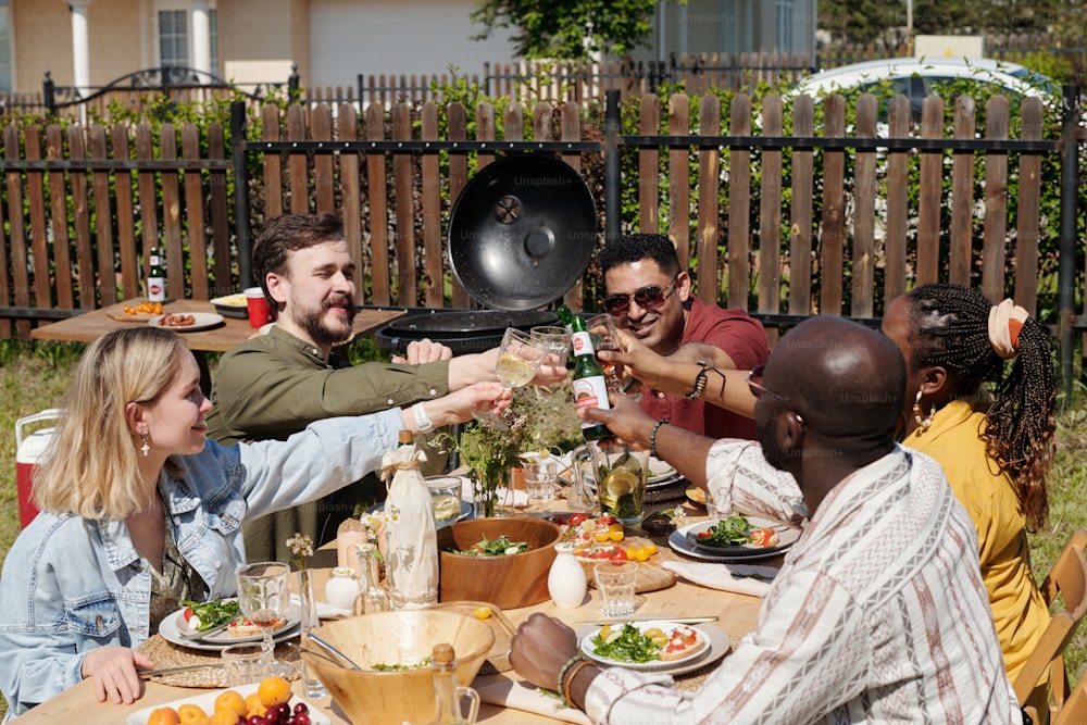 Group of young interracial friends with drinks toasting during outdoor dinner or party over table served by homemade food and beverages
