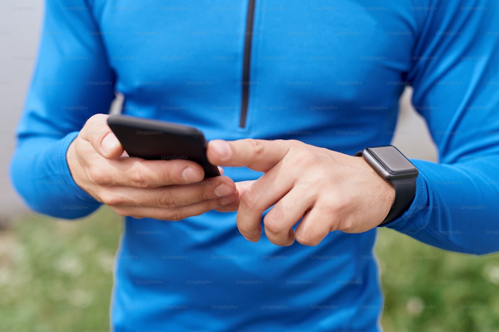 Nahaufnahme eines bärtigen Sportlers mittleren Alters, der seine Smartwatch mit dem Smartphone überprüft, synchronisiert, im Sommer im Freien trainiert, an einem düsteren Tag mit malerischer Aussicht, im blauen Hemd