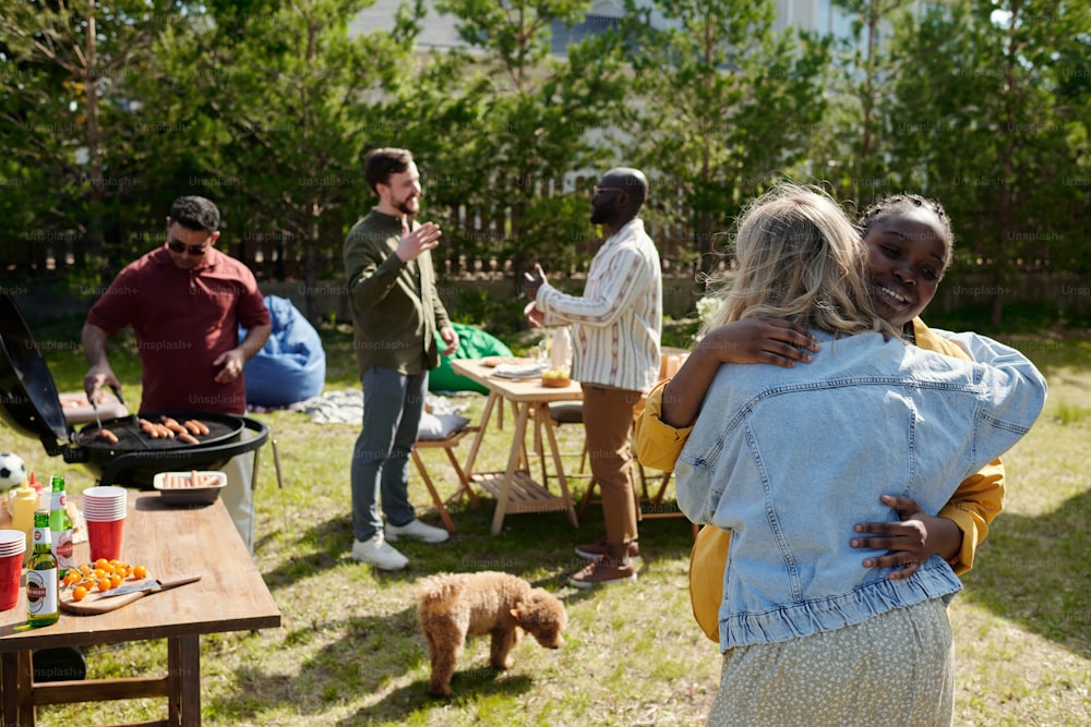 Happy young intercultural women embracing each other against two men having discussion and their friend making barbecue on grill