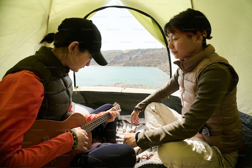 Two modern young Asian women sitting in camp tent at quarry lake shore relaxing and playing ukulele