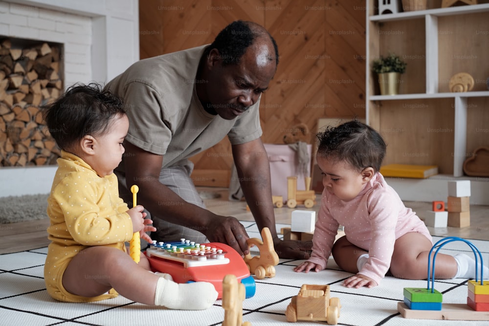 Padre afroamericano moderno sentado en el suelo en la sala de estar de su casa jugando con sus encantadores bebés gemelos