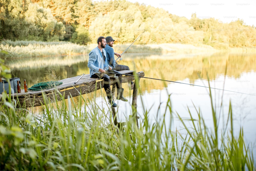 Dos amigos vestidos con camisas azules pescando juntos con red y caña sentados en el muelle de madera durante la luz de la mañana en el lago